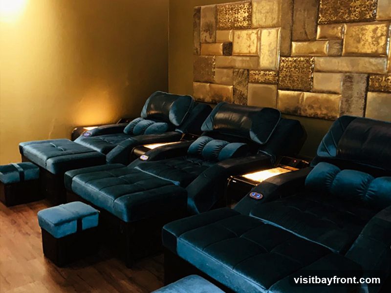 A row of plush, dark teal massage chairs with footrests are arranged in a dimly lit room. The chairs have built-in controls and illuminated side tables. The wall behind the chairs is decorated with a textured, metallic gold paneling.