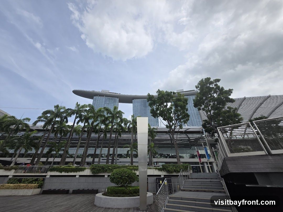 A wide-angle shot of the Marina Bay Sands complex in Singapore under a cloudy sky. The iconic three towers topped with the ship-like structure are visible in the background. In the foreground, there are palm trees, lush greenery, and modern architectural elements including stairs and walkways. The overall scene depicts a large, contemporary urban landscape with a focus on impressive architecture and landscaping.