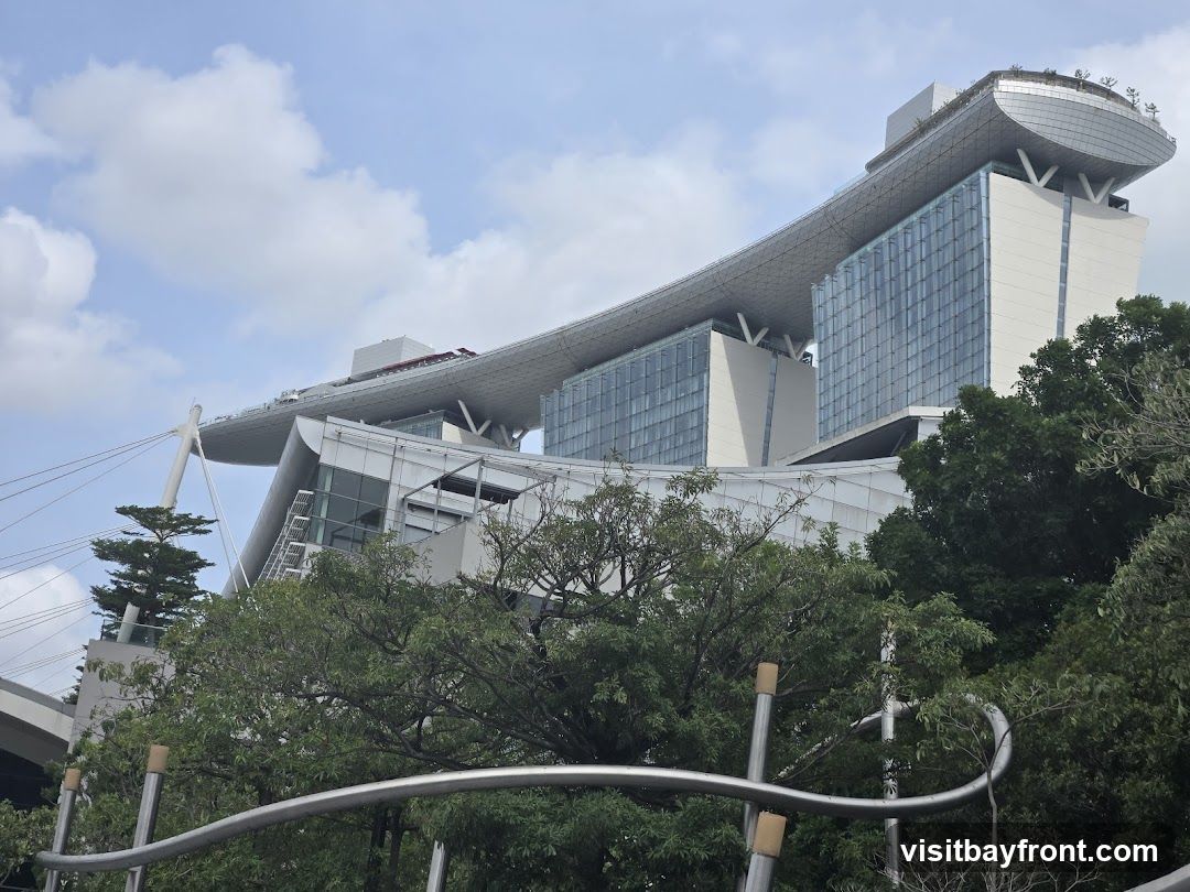 A low-angle shot of a modern architectural complex featuring a distinctive sky bridge connecting two towers. The building has a sleek, contemporary design with glass facades and white structural elements. Lush green trees partially obscure the lower levels of the structure, contrasting with the clear blue sky and scattered clouds above.
