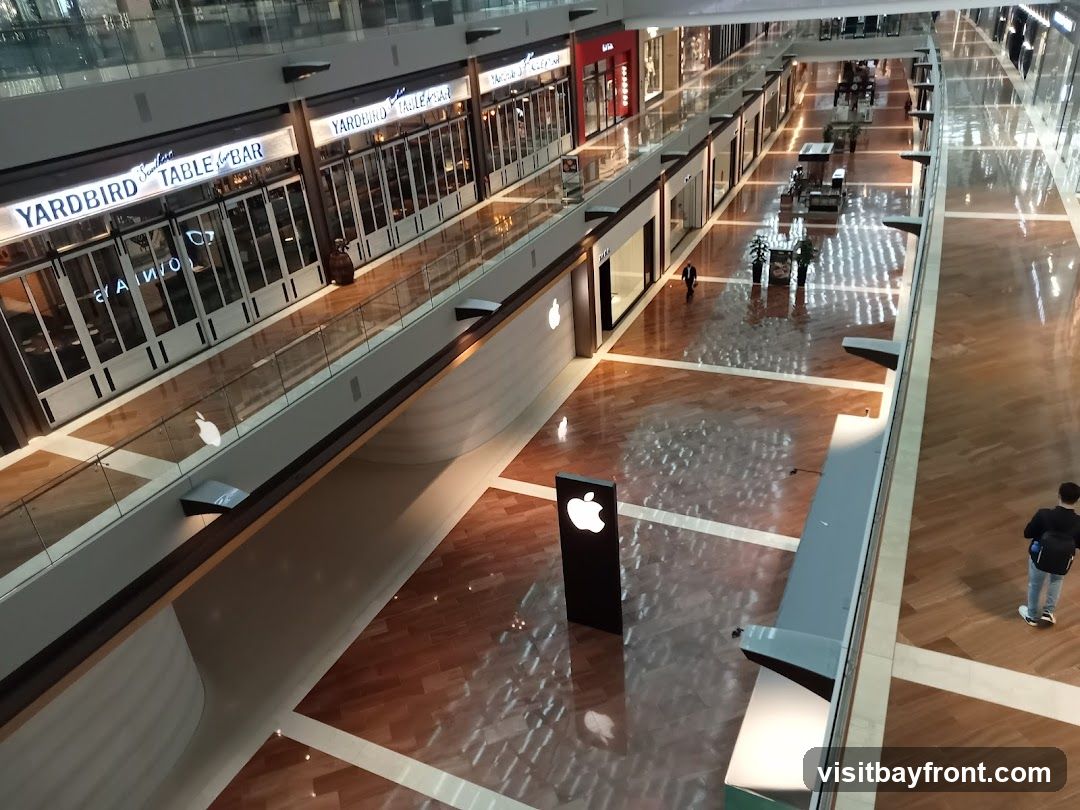 An aerial view of a modern indoor shopping mall. The image shows multiple levels with various storefronts lining the walkways. On the left, a restaurant with the sign "YARD BIRD TABLE & BAR" is visible. In the center, an Apple Store is indicated by prominent Apple logos displayed on signage and on the store's facade. People are seen walking through the mall, and the polished wooden floors reflect the overhead lighting, creating a bright and spacious atmosphere.