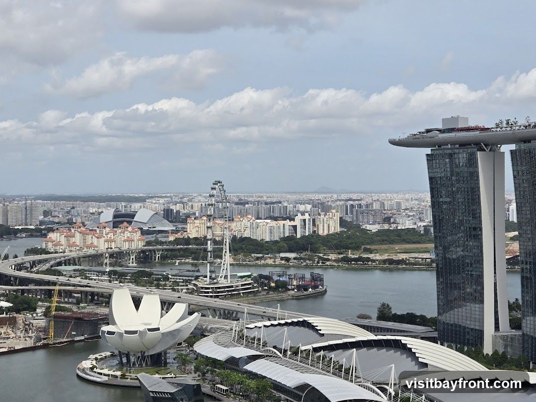 A panoramic view of the Singapore skyline on a cloudy day. Prominent structures include the Marina Bay Sands hotel with its iconic rooftop infinity pool, the ArtScience Museum resembling a lotus flower, a Ferris wheel, and a stadium. The cityscape extends to the horizon, with numerous buildings and infrastructure visible.