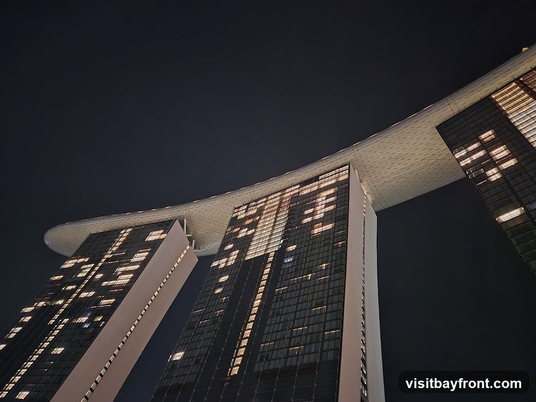 A low-angle, night-time shot of the iconic Marina Bay Sands hotel in Singapore. The three towers are connected by a large, curved rooftop structure. Many windows on the towers are illuminated, creating a grid-like pattern of light against the dark sky.