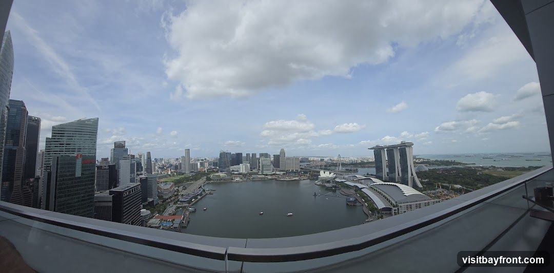 A panoramic view of the Singapore skyline, featuring numerous skyscrapers and the Marina Bay Sands hotel. The image is taken from a high vantage point, likely a rooftop or balcony, with a railing in the foreground. The sky is partly cloudy, with a mix of blue sky and white clouds. Several boats are visible on the water.
