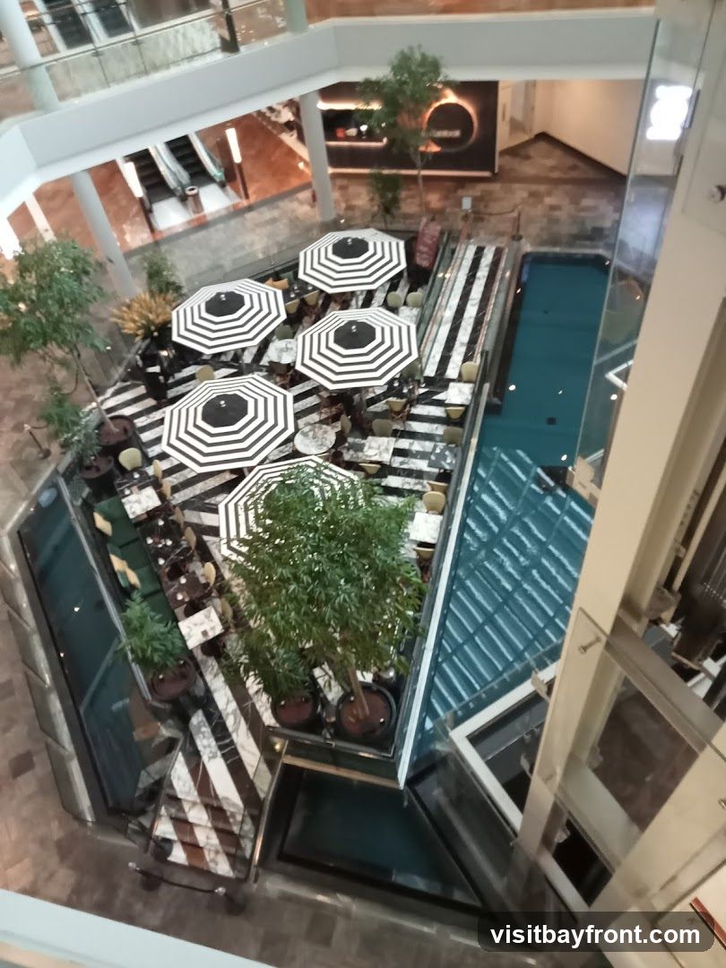An aerial view of an outdoor seating area within a shopping mall. The area features black and white striped umbrellas over tables and chairs, surrounded by potted plants and a reflective pool. Escalators and other mall levels are visible in the background.