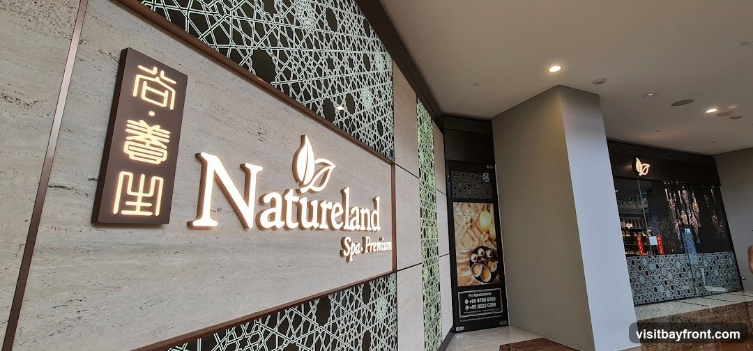 A close-up view of the Natureland Spa Premium storefront. The signage features the spa's name in illuminated white lettering, accompanied by a leaf logo. To the left of the main sign, there is a vertical panel with Chinese characters, also illuminated. The facade is decorated with a patterned screen. To the right, the entrance to the spa is visible, with glass doors and a dark frame. Inside, shelves with products can be seen. A poster with contact information and images of spa treatments is displayed next to the entrance.