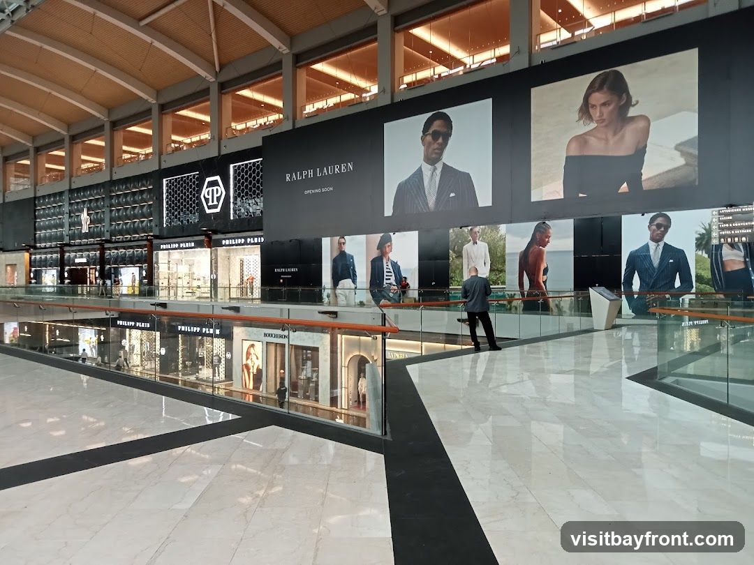A wide shot of a modern shopping mall interior. Large digital screens display advertisements for fashion brands, including Ralph Lauren and Philipp Plein. The screens feature models in stylish clothing. Below the screens, storefronts of various luxury brands are visible, with their names like Philipp Plein and Boucheron displayed. The mall has a high ceiling with exposed beams and lighting. The floor is polished marble, reflecting the lights and advertisements. A few people are seen walking around the mall.