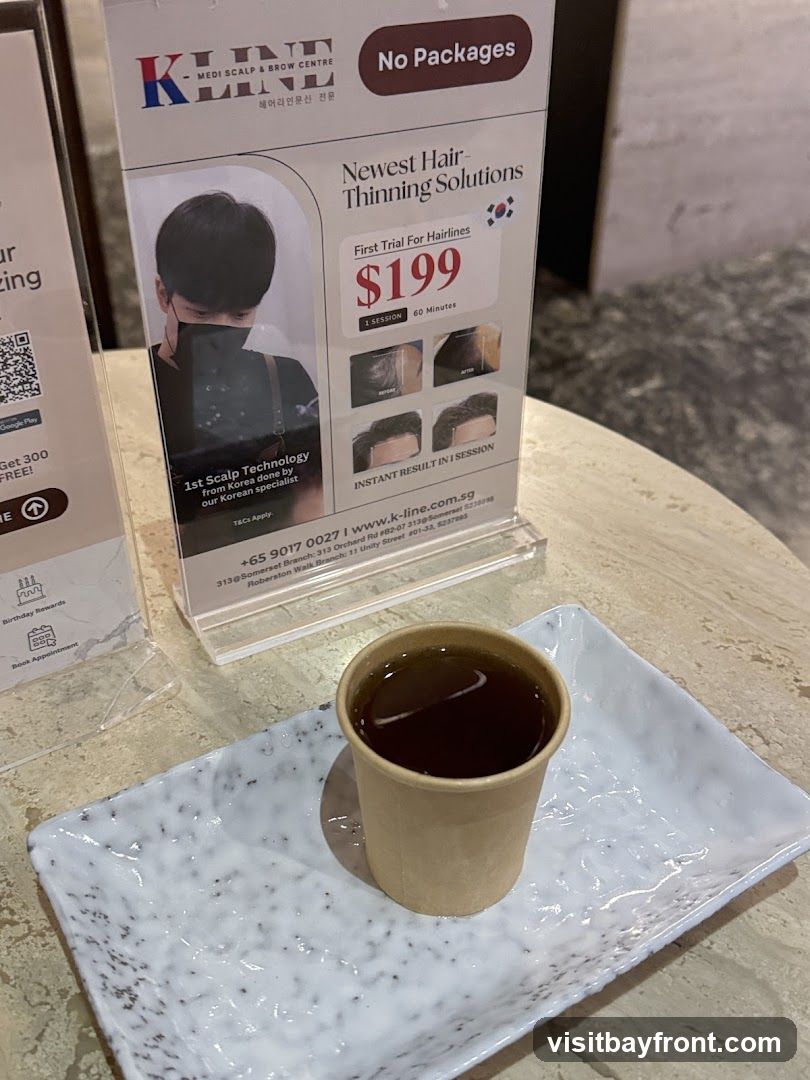 A promotional display for K-Line Medi Scalp & Brow Centre, advertising their newest hair-thinning solutions. The display features a trial offer for hairlines at $199 for one 60-minute session, showcasing before and after images. It highlights "1st Scalp Technology from Korea done by our Korean specialist." Below the main advertisement, contact information and branch addresses are provided. A cup of dark liquid, likely tea, is served on a speckled white tray in the foreground.