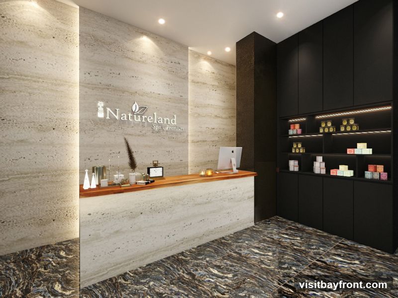 A modern spa reception area with a wooden counter, a computer, and decorative items. Behind the counter, a wall features the spa's logo, "Natureland Spa, Premium." To the right, black cabinets with illuminated shelves display various spa products. The floor has a marbled pattern.