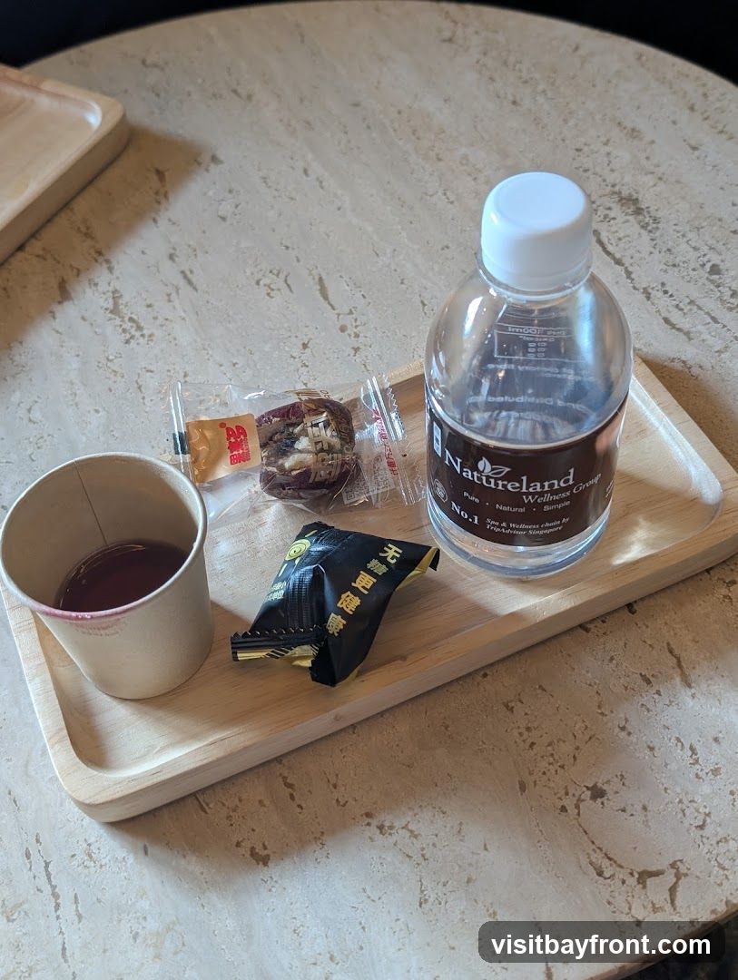 A wooden tray holds a cup of dark liquid with lipstick marks on the rim, two individually wrapped snacks, and a bottle of Natureland Wellness Group water. The water bottle label indicates it is from a spa and wellness chain in Singapore.