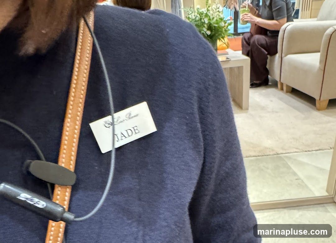 A close-up of a person wearing a dark blue sweater. A name tag is pinned to the sweater, reading "Loro Piana JADE". A brown leather strap and black earbuds are also visible. In the background, a person is seated in a chair, holding a green drink, and there is a table with flowers.