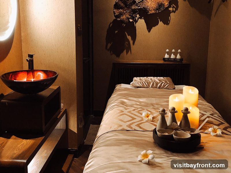 A dimly lit spa room with a massage bed, a sink, and decorative elements. The massage bed is covered with a beige and gold patterned sheet and a satin pillow. Several large candles are lit on the bed, along with small bottles and bowls, possibly for massage oils or scrubs. White plumeria flowers are scattered around the bed. To the left, a dark wooden vanity holds a red glass sink with a modern faucet. The walls are textured in a warm, earthy tone, and a decorative piece hangs on the wall above a dark wooden cabinet.