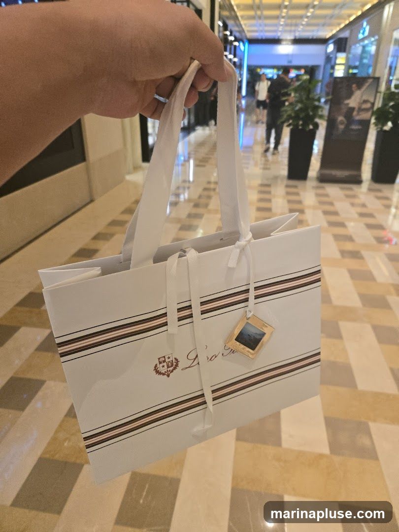 A hand holds a white shopping bag with a logo and striped design. The bag has white ribbon handles and a small decorative tag. The background shows a blurred indoor shopping mall with tiled flooring and other shoppers.