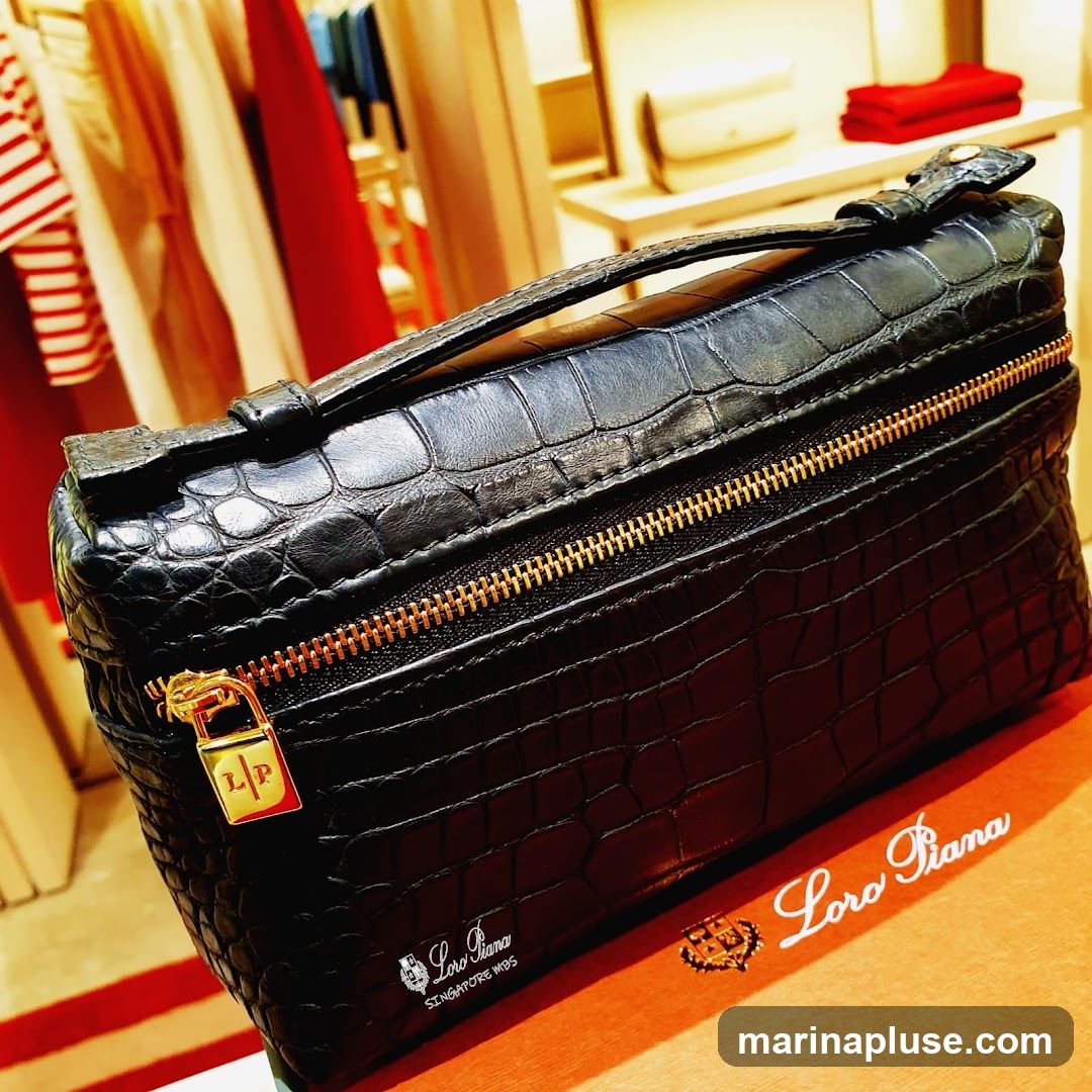 A close-up shot of a black crocodile leather bag with gold hardware. The bag has a top handle and a zippered closure. A small padlock with the initials 'LP' is attached to the zipper. The Loro Piana logo is visible on the bag and on an orange box in the foreground. In the background, clothing items are displayed on racks.