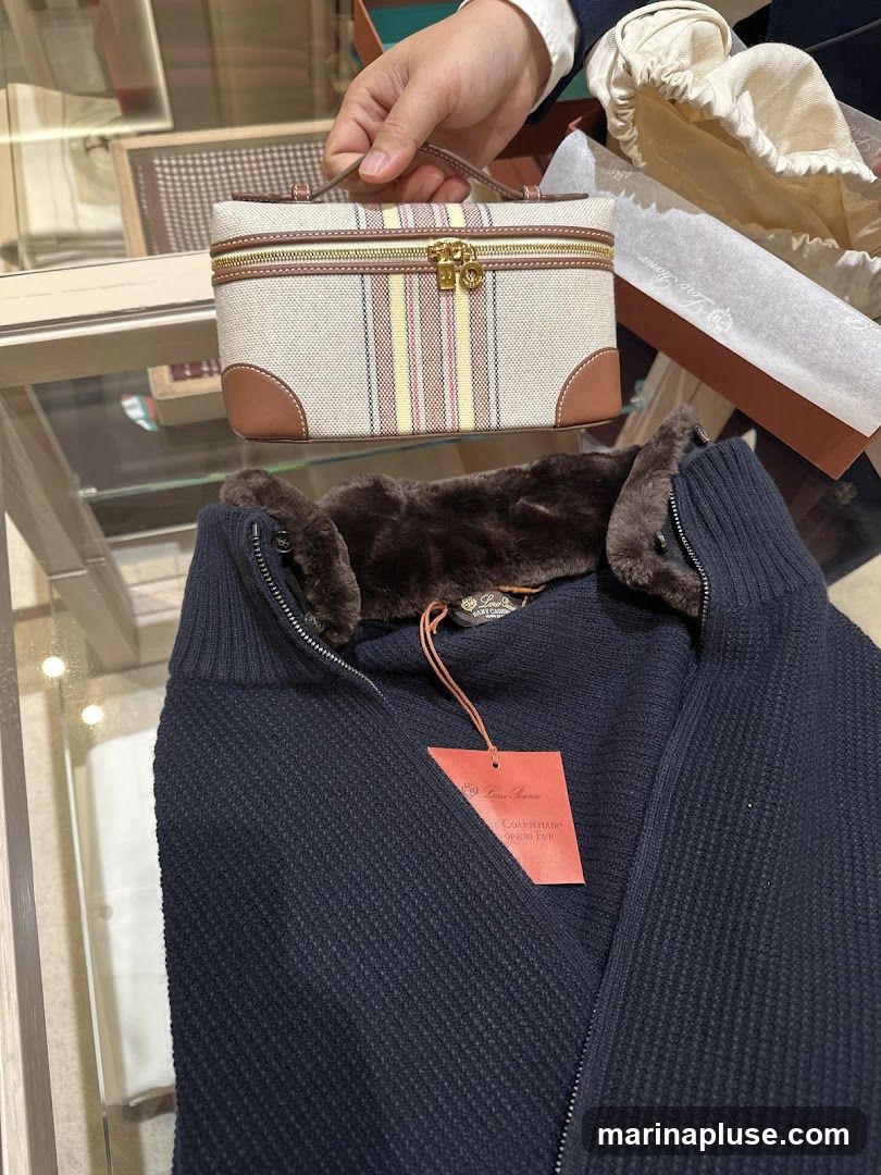 A close-up view of a navy blue knitted sweater with a dark brown fur collar, featuring a Loro Piana tag. In the background, a person's hand holds a small, striped vanity case with leather trim and gold hardware. The items appear to be displayed on a glass surface, possibly in a retail setting.