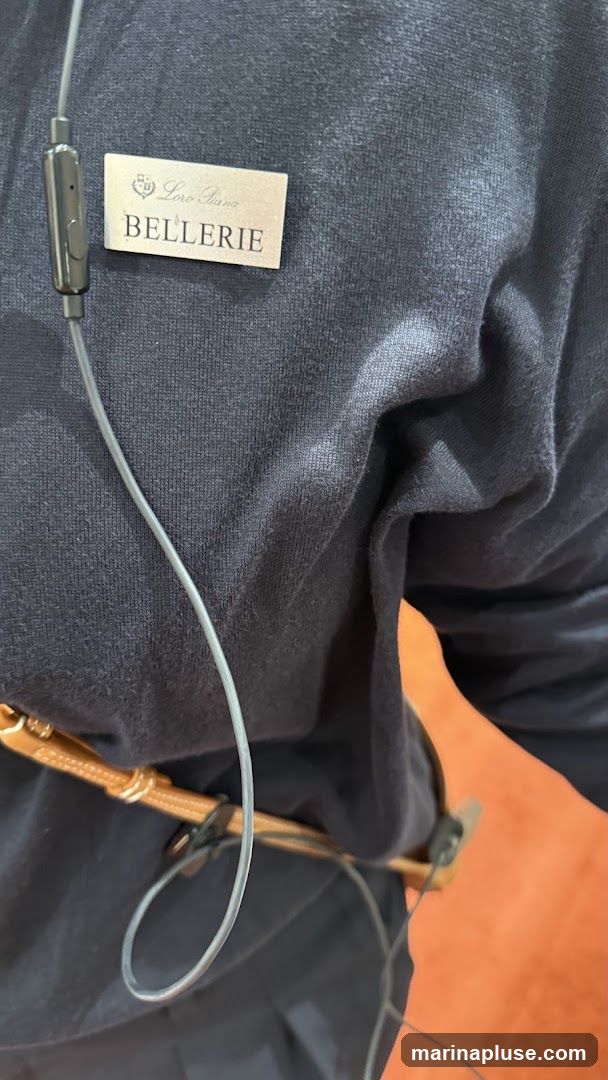 A close-up of a dark blue garment with a name tag attached. The name tag is silver and reads "Loro Piana BELLERIE" with a crest above the text. A grey earphone wire is also visible, draped over the garment.