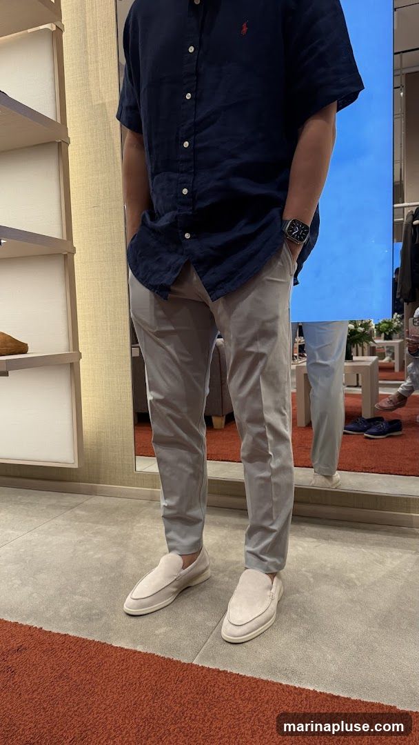A person stands in a store, wearing a navy blue short-sleeved shirt with a red logo on the chest, light grey pants, and light-colored loafers. The person is wearing a watch on their left wrist. The background shows shelves with merchandise, a large mirror reflecting the scene, and other shoppers.