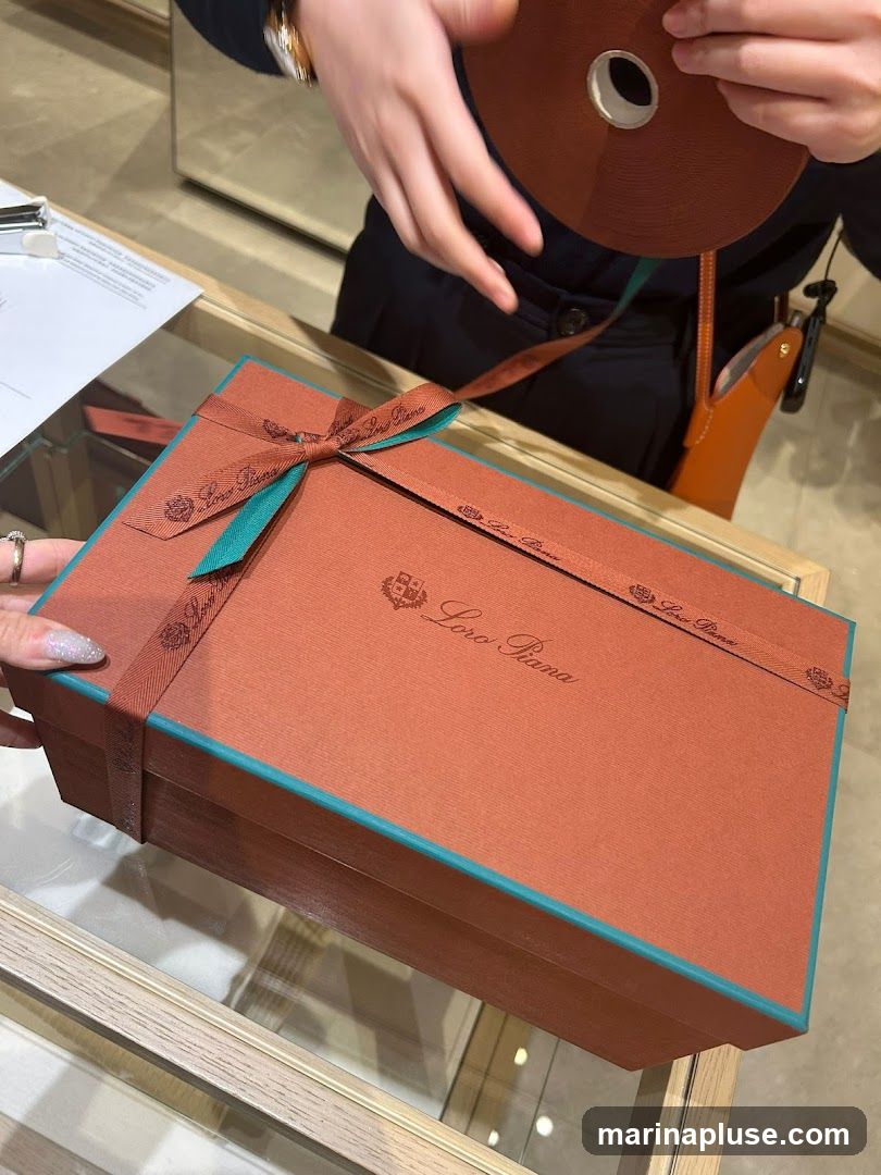 A close-up shot of a Loro Piana gift box, tied with a brown and teal ribbon. The box is orange and has the Loro Piana logo embossed on it. A person's hands are visible, one holding a roll of ribbon and the other adjusting the bow on the box. The scene appears to be inside a store, with a glass display case in the foreground.
