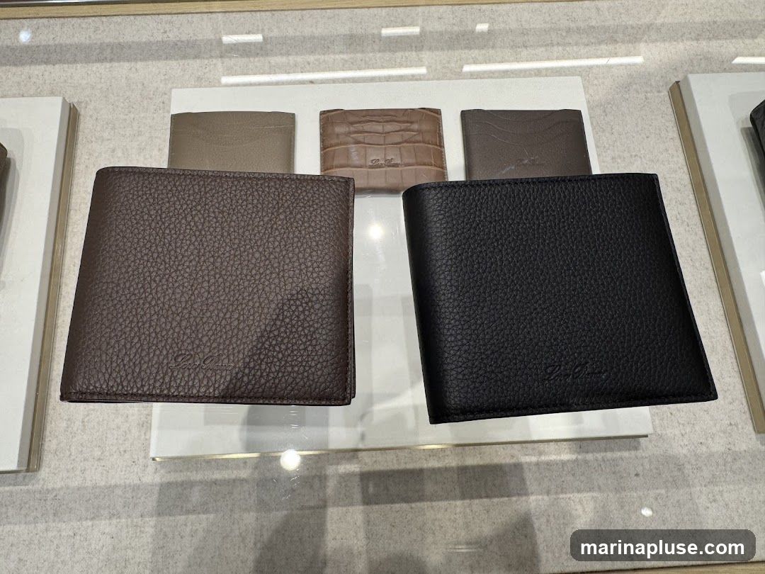 Two textured leather wallets, one brown and one black, are displayed prominently in the foreground. Behind them, several other wallets are visible, including one with a crocodile-embossed pattern and others in muted tones. The wallets appear to be laid out on a display shelf or in a store window.