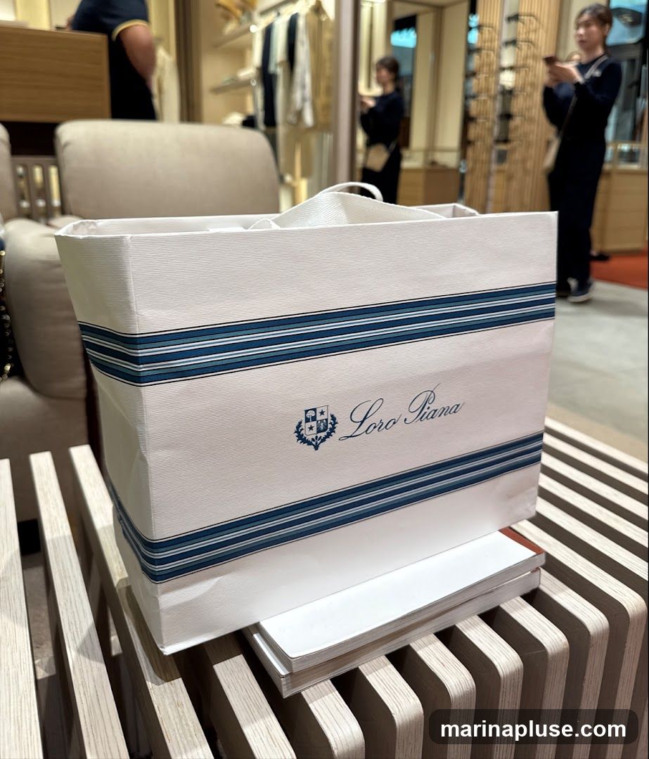 A white shopping bag with blue stripes and the Loro Piana logo is placed on a wooden surface. In the background, a store interior with clothing racks and customers is visible.