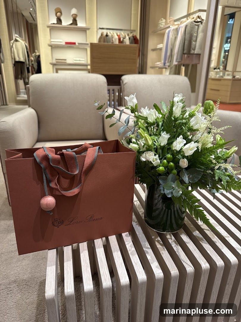A Loro Piana shopping bag sits on a slatted wooden table next to a vase of white flowers. The bag is a muted rose color with the Loro Piana logo printed in the center. The handles are a striped ribbon. The background shows a well-lit retail store interior with clothing displays and seating areas.