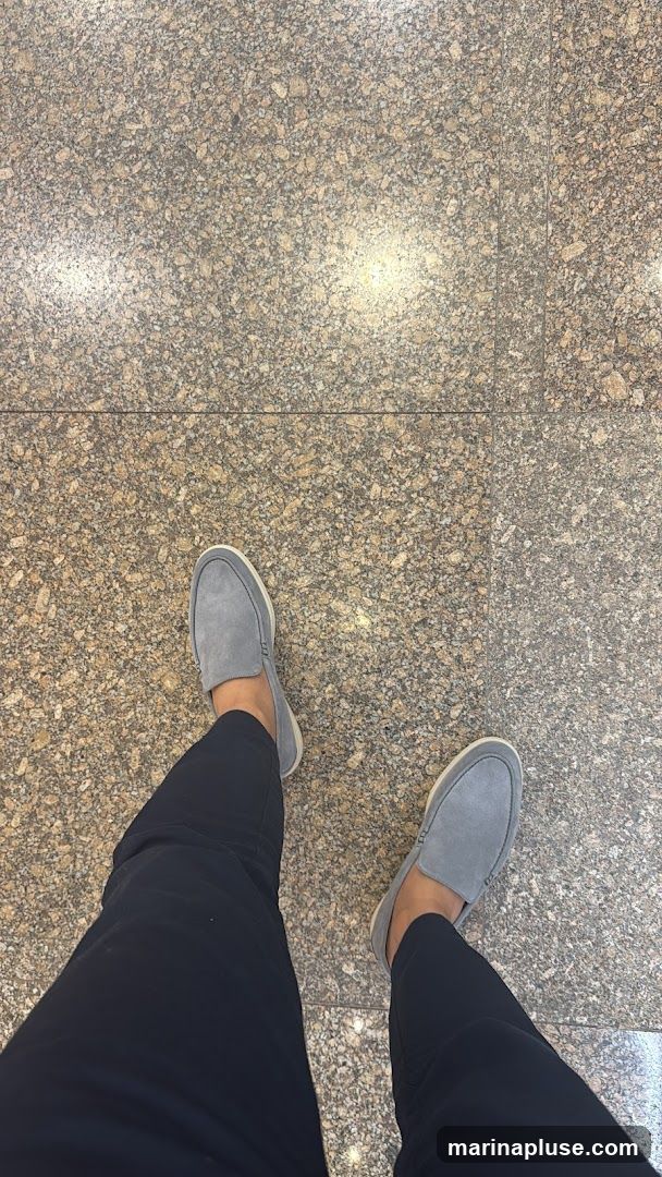 A first-person perspective shot of a person's feet wearing gray suede loafers and black pants. The person is standing on a floor tiled with speckled granite.
