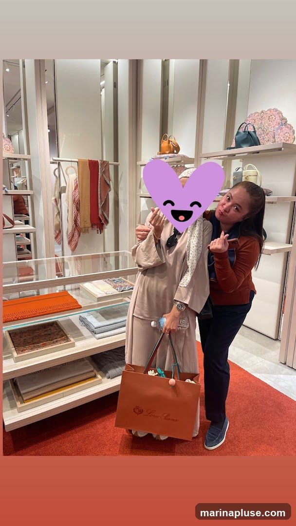 Two women are posing for a photo inside a store. The woman on the left is wearing a beige abaya and a patterned scarf, with a purple heart emoji covering her face. She is holding a shopping bag with the logo "Loro Piana" visible. The woman on the right is wearing a rust-colored long-sleeved shirt and dark pants. The store interior features shelves displaying scarves, bags, and other accessories. The shelves are made of light-colored wood and have white vertical accents. The floor is covered with a red carpet.