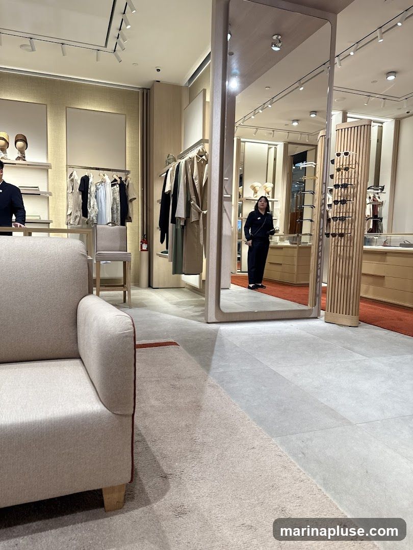 A well-lit retail store interior with clothing racks, display shelves, and a large mirror. A person in dark clothing stands near the mirror, and another person in a dark uniform stands behind a counter on the left. A sofa is visible in the foreground. The store appears to be selling apparel and accessories.