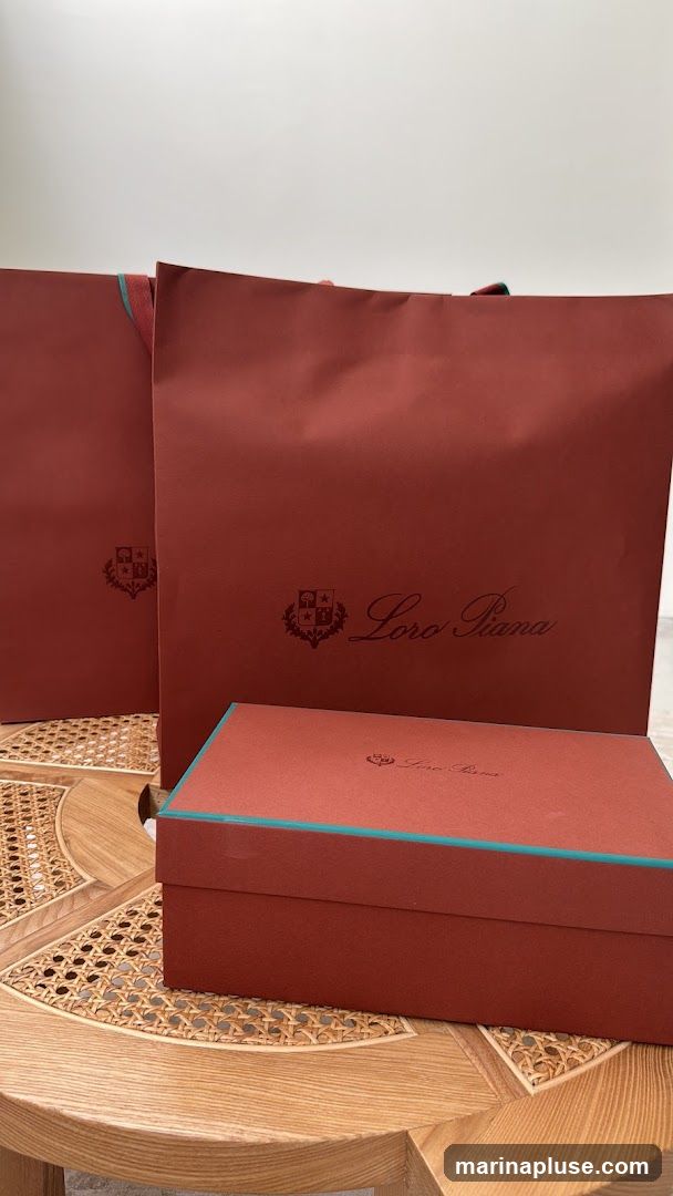 Two rust-colored shopping bags and a matching box with teal trim are displayed on a round wooden table with a woven cane inlay. The bags and box feature the Loro Piana logo and crest.