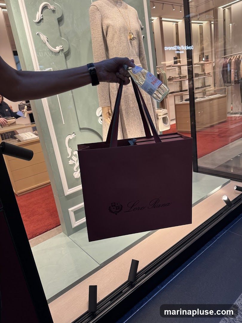 A person's arm holds a shopping bag with the Loro Piana logo and a bottle of Acqua Panna water in front of a store window display. The display features a mannequin in a beige dress and clothing racks with apparel.
