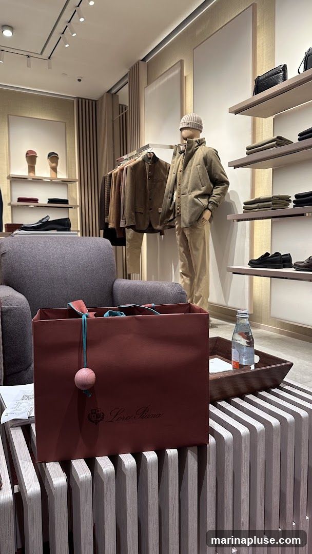A Loro Piana shopping bag is prominently displayed on a slatted wooden surface in a well-lit retail store. The bag is a deep red with the Loro Piana logo in script. Next to the bag, on a wooden tray, is a bottle of water and a small white card. In the background, mannequins are dressed in stylish outerwear and trousers, with racks of clothing and shelves displaying shoes and accessories. The store interior features neutral tones and modern design elements.