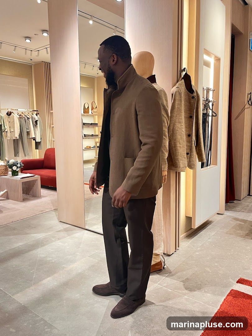 A man stands in a well-lit clothing store, looking into a large mirror. He is wearing a light brown jacket and dark brown trousers. In the background, clothing racks with various garments, a red sofa, and mannequins displaying outfits are visible. The store appears to be a high-end boutique with a minimalist and modern aesthetic.