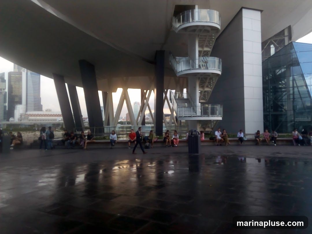 A group of people are sitting on a ledge under a large, modern architectural structure. The structure features a prominent staircase with circular landings and a glass-fronted section. In the background, a cityscape with tall buildings is visible. The ground in the foreground is wet and reflective.