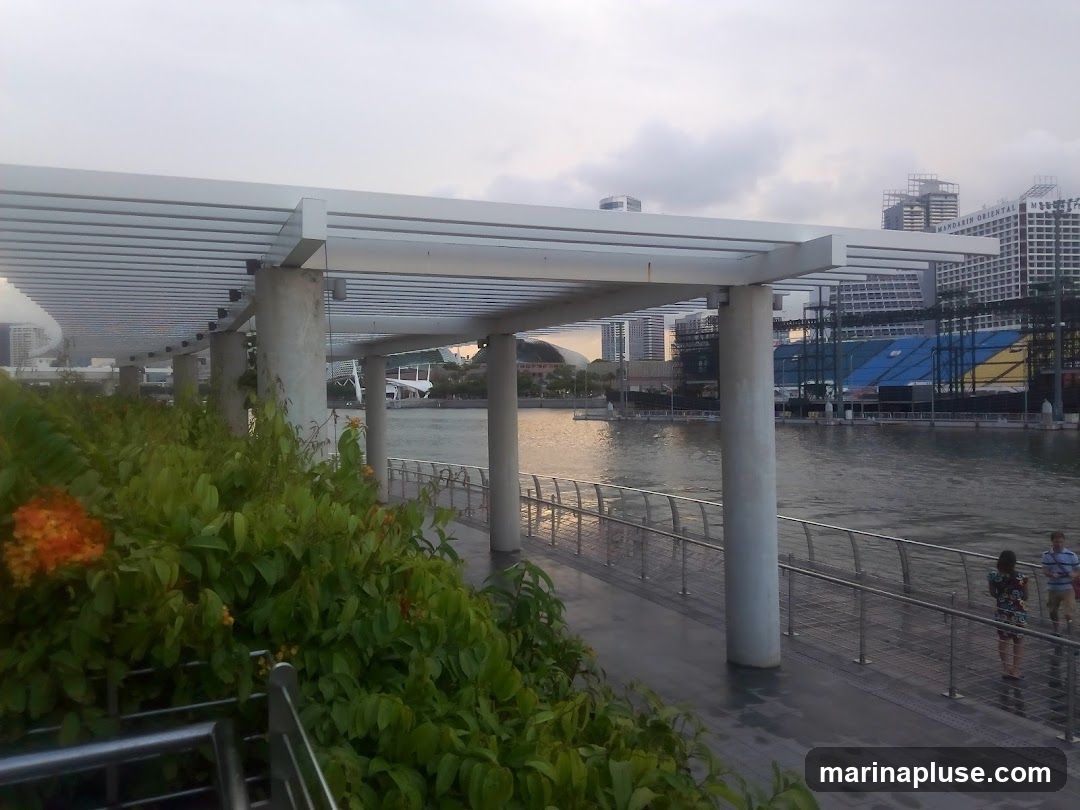 A waterfront promenade with lush green foliage and orange flowers in the foreground. A modern white canopy structure supported by concrete pillars extends along the walkway. In the background, a body of water reflects the overcast sky, and a cityscape with numerous buildings, including the Mandarin Oriental, is visible. A large structure with blue and yellow sheeting is under construction or renovation on the water. Two people are walking on the promenade.