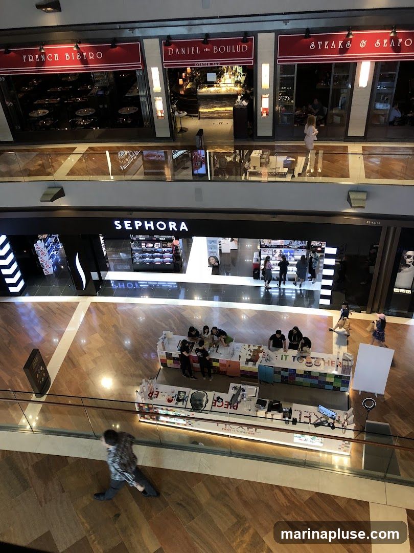 An overhead view of a shopping mall interior. On the upper level, there are three distinct storefronts: "FRENCH BISTRO", "DANIEL BOULUD OYSTER BAR", and "STEAKS & SEAFOOD". Below, a Sephora store is visible with its prominent signage. In the center of the lower level, a long, colorful kiosk with various products and displays is set up, with several people interacting with it. A person is walking in the foreground on the polished wooden floor.
