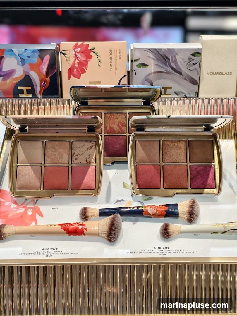 A close-up view of an Hourglass cosmetics display featuring several Ambient Lighting Edit palettes and brushes. The palettes are open, showing various shades of blush and highlighter. Two makeup brushes with floral designs are laid out in front of the palettes. The packaging and products are arranged on a display shelf with a ribbed, metallic finish.