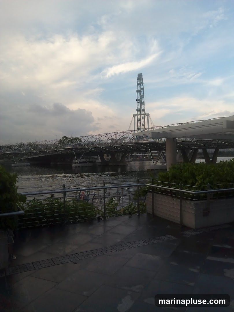 A view of a waterfront promenade with a modern bridge and a tall tower in the background. The sky is cloudy with patches of blue. The promenade is paved with dark tiles, and there are metal railings and planters with greenery. The water is calm.