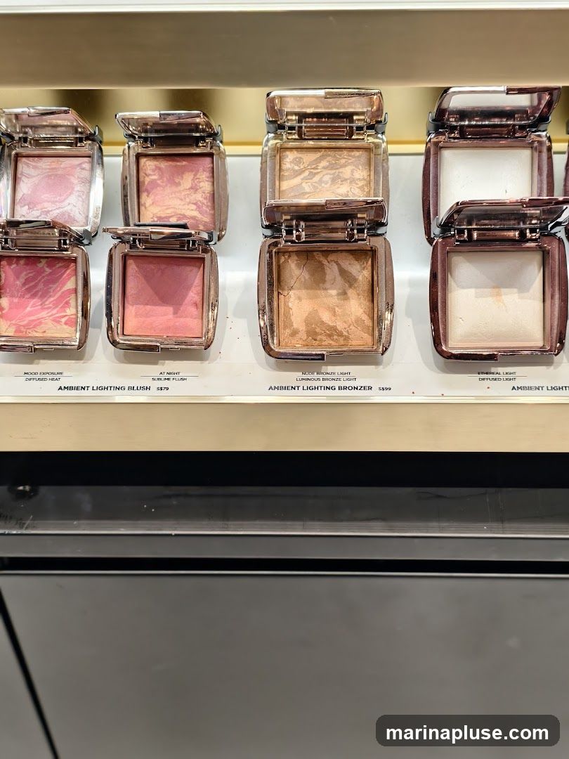A display of several Hourglass Ambient Lighting products, including blushes and bronzers, arranged on a shelf. The products are open, showing their marbled powders. Labels with product names and prices are visible below each item.