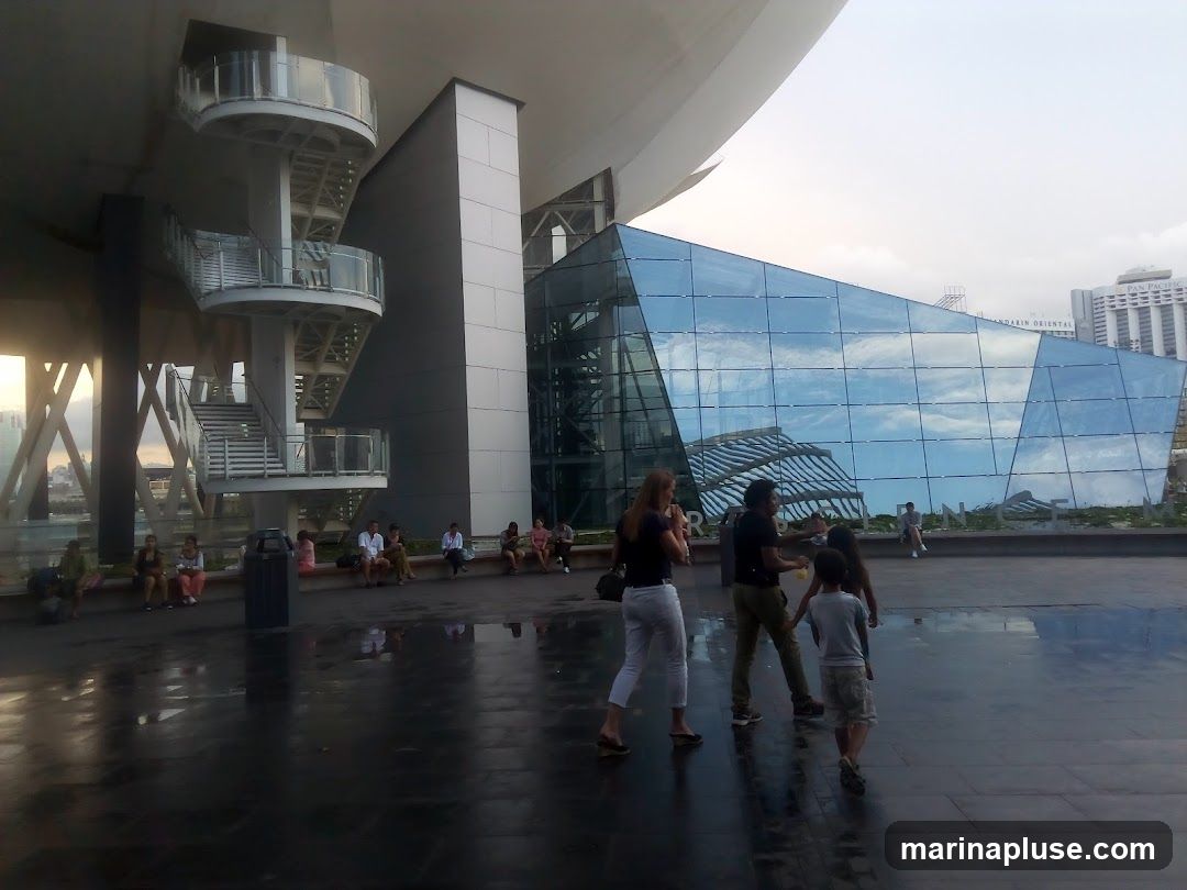A wide shot of a modern building with a distinctive glass facade, reflecting the sky and surrounding structures. A spiral staircase is visible on the left. Several people are walking and sitting in the foreground and midground, enjoying the outdoor space. The overall scene suggests a public area or a cultural institution.