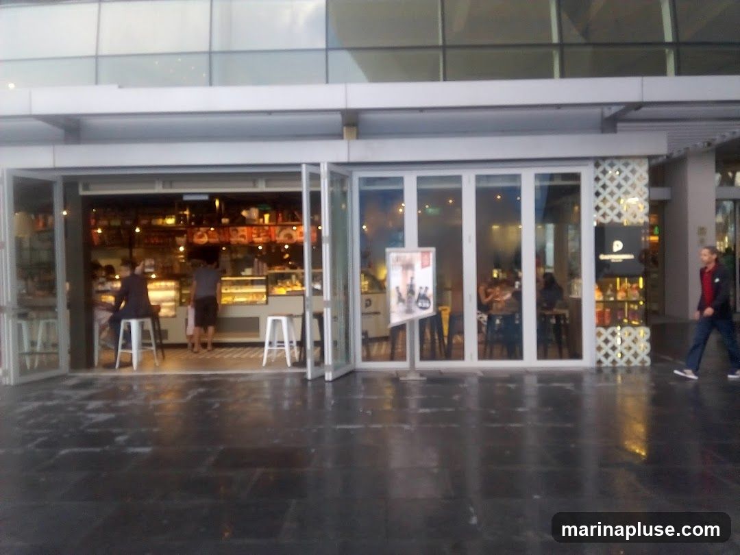 A wide shot of a cafe's exterior with large glass doors and windows. Inside, people are seated at tables and at a counter. Display cases show pastries and other food items. A sign with a logo is visible on the right side of the entrance. A man walks past the cafe on the right.