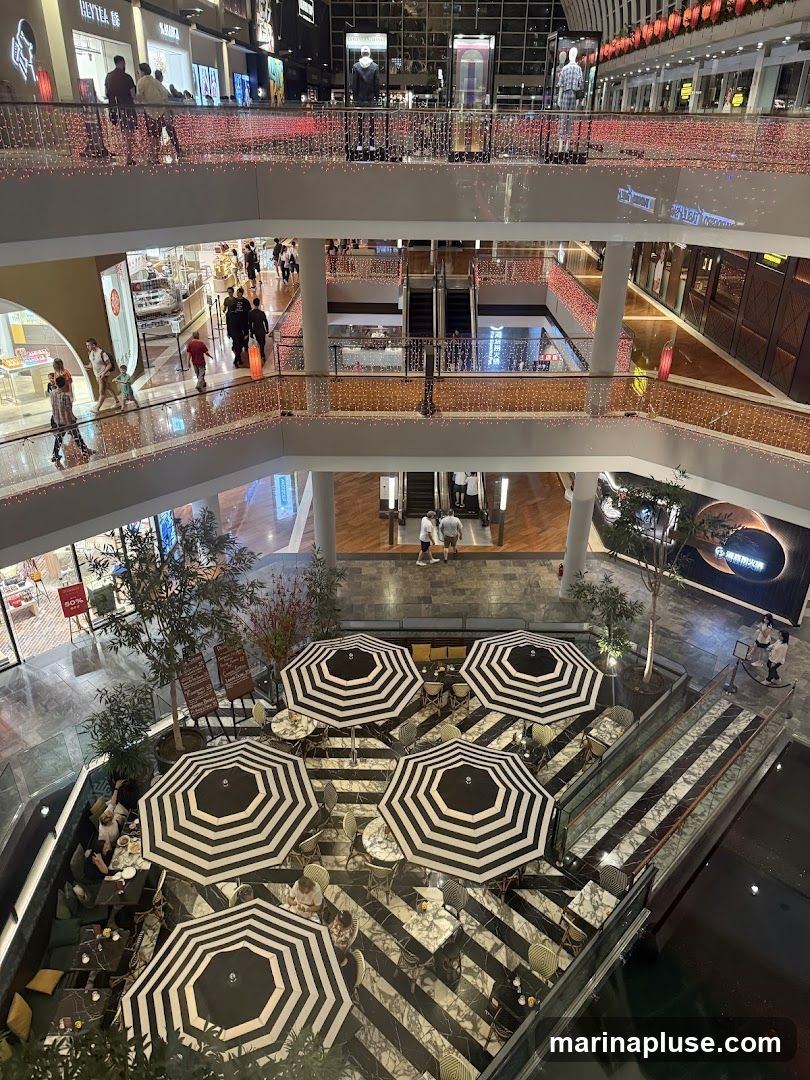 An aerial view of a multi-level shopping mall. The upper levels show various shops and escalators, decorated with strings of red lights. The lower level features an outdoor seating area with black and white striped umbrellas covering tables and chairs. Several people are seated at the tables, and others are walking on the upper levels.