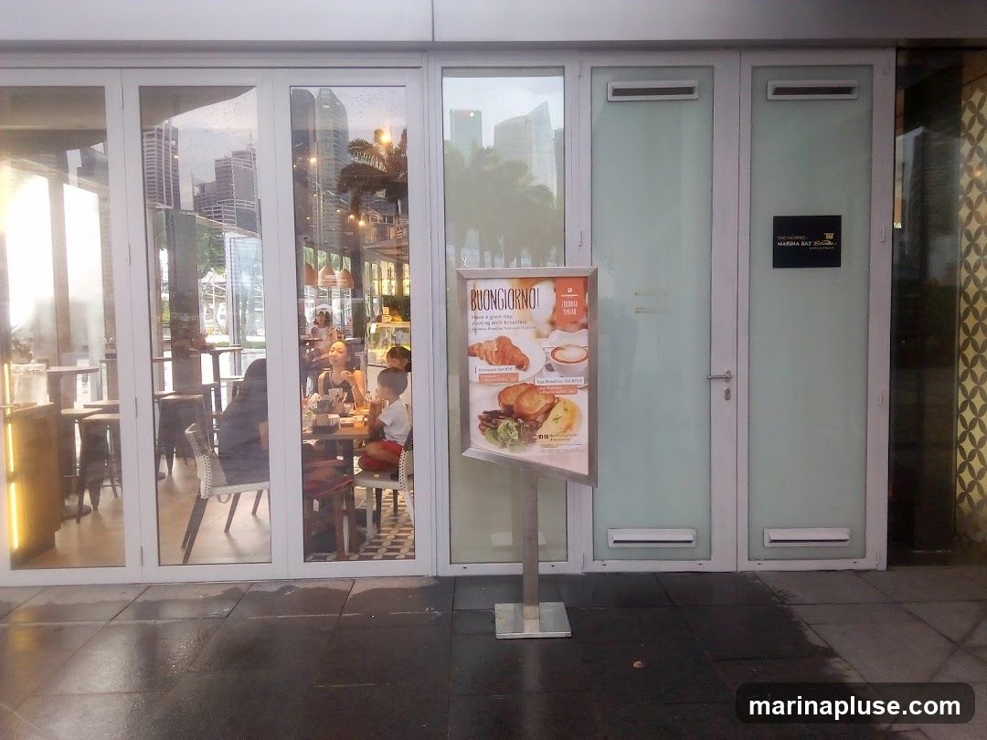 A cafe with large glass windows and doors, showing people seated inside at tables. A menu board is placed outside the entrance, displaying breakfast items. The cafe has a modern design with clean lines.