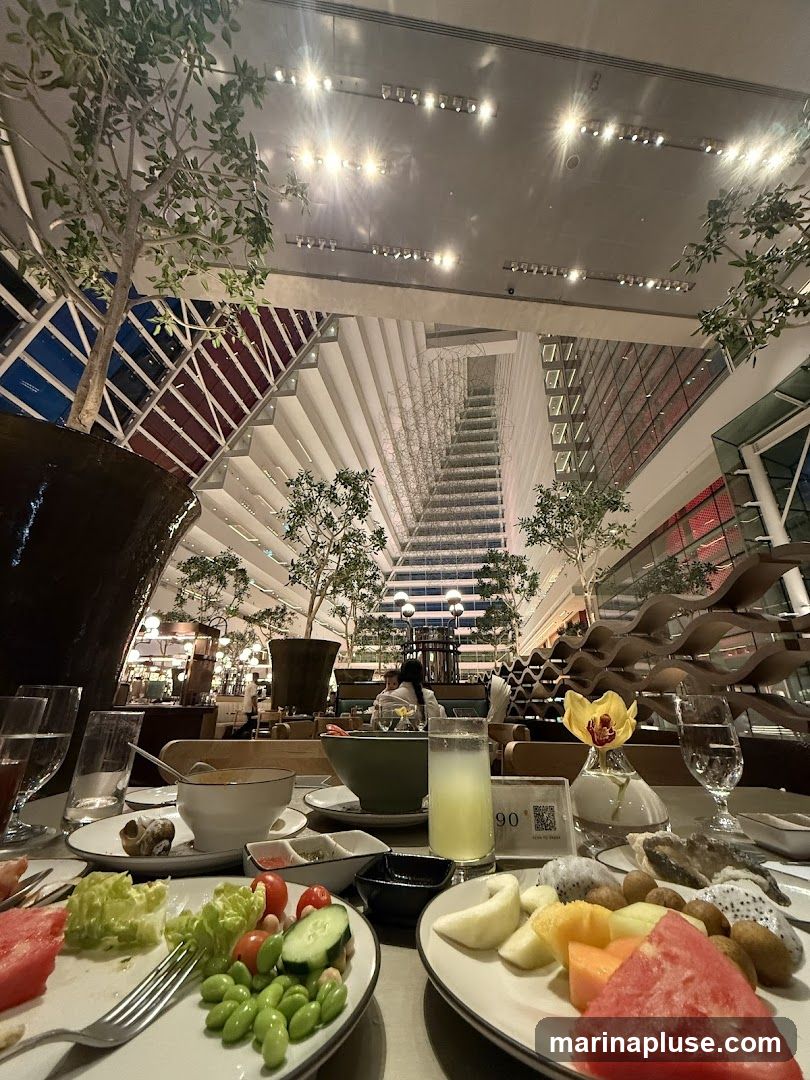 A close-up view of a buffet table laden with various dishes, including a fresh salad with lettuce, tomatoes, cucumbers, and edamame, a plate of sliced fruits such as watermelon, papaya, and dragon fruit, and a glass of yellow beverage. In the background, the interior of a modern building with a grand staircase and decorative trees is visible.