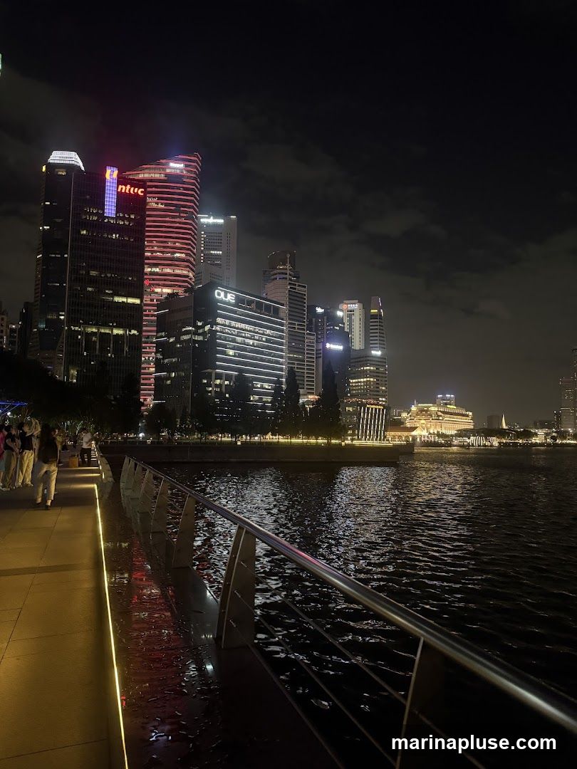A nighttime view of a city skyline with illuminated skyscrapers and a waterfront promenade. The water reflects the city lights, creating a shimmering effect. Several people are walking along the promenade.