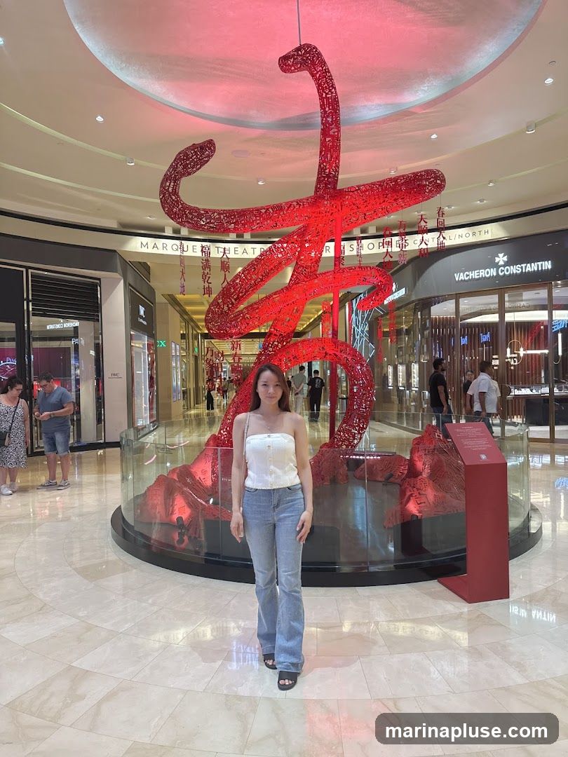 A woman stands in the center of a mall, posing in front of a large, intricate red sculpture. The sculpture appears to be made of intertwined metal or wire, forming abstract shapes. In the background, various shops are visible, including one with a prominent Vacheron Constantin sign. The mall's interior features a polished floor and a curved ceiling with lighting. Other shoppers can be seen in the distance.