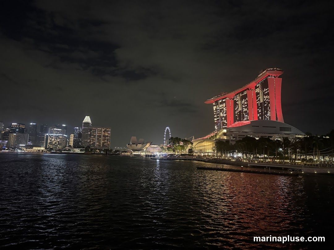 A nighttime view of the Singapore skyline, featuring the iconic Marina Bay Sands hotel illuminated in red, along with other skyscrapers and a Ferris wheel. The water in the foreground reflects the city lights.