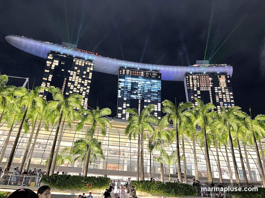 A nighttime view of the Marina Bay Sands hotel in Singapore, with its iconic three towers topped by a ship-like structure. Lights illuminate the building and a laser show projects beams into the dark sky. Palm trees line the foreground, with a crowd of people visible on a walkway below.