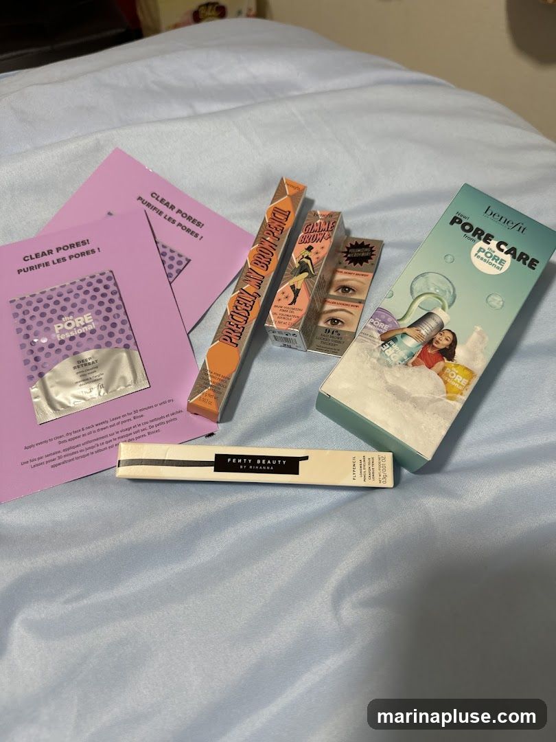 A collection of Benefit and Fenty Beauty makeup products, including brow pencils, pore care items, and eyeliner, laid out on a light blue fabric surface.