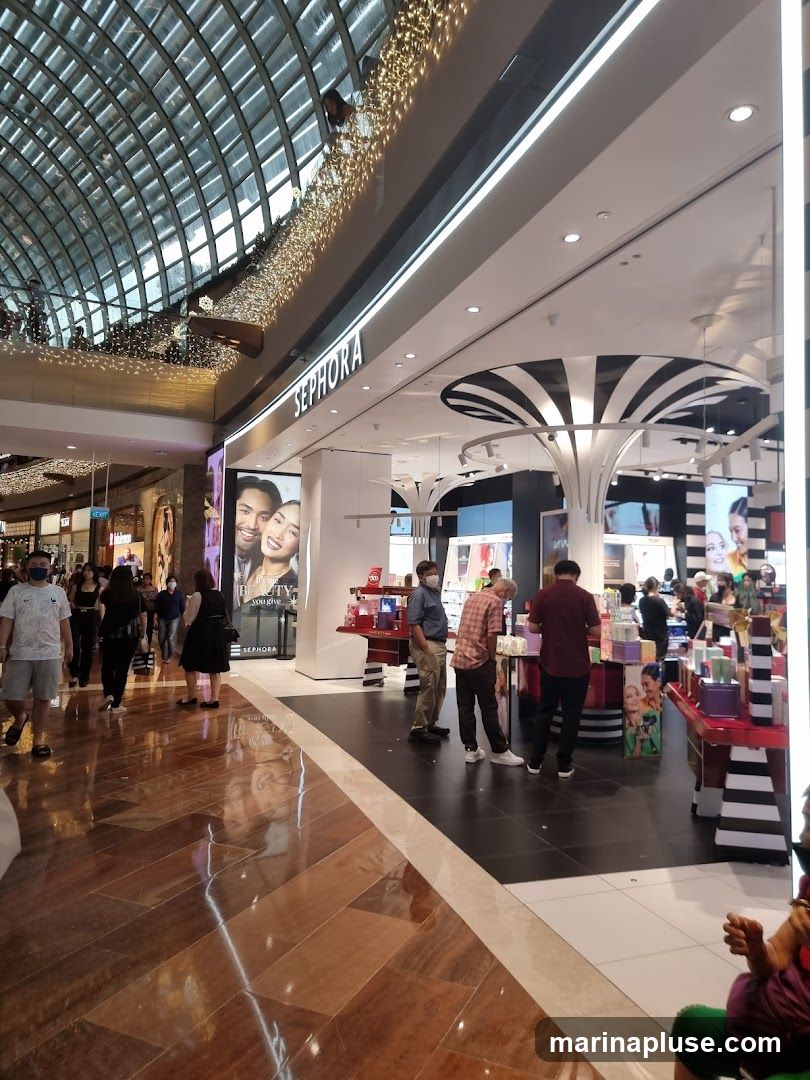 The exterior of a Sephora store is visible, with the brand name prominently displayed above the entrance. The store is located within a mall, indicated by the arched ceiling with decorative lights and the polished floor. Several customers are seen browsing inside and walking past the entrance. The interior features displays of beauty products and promotional imagery.
