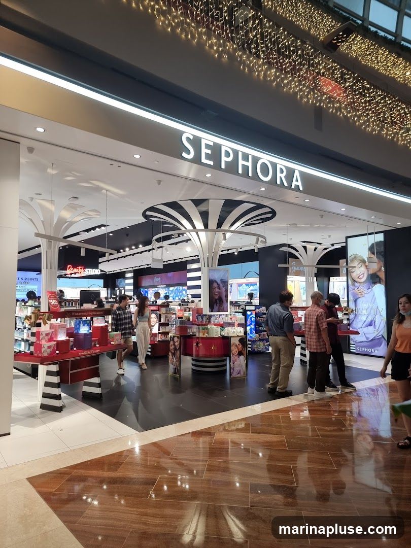The exterior of a Sephora store with the brand name prominently displayed above the entrance. Inside, various beauty products are displayed on shelves and stands. Several customers are browsing the merchandise. The store is illuminated by bright lights and decorated with festive string lights overhead.