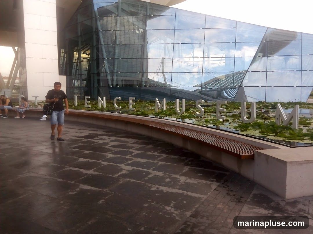 A man walks past a modern building with a glass facade. Large metal letters spelling out "SCIENCE MUSEUM" are visible in front of the building, with a water feature and lily pads below. Several people are seated on a bench in the background.